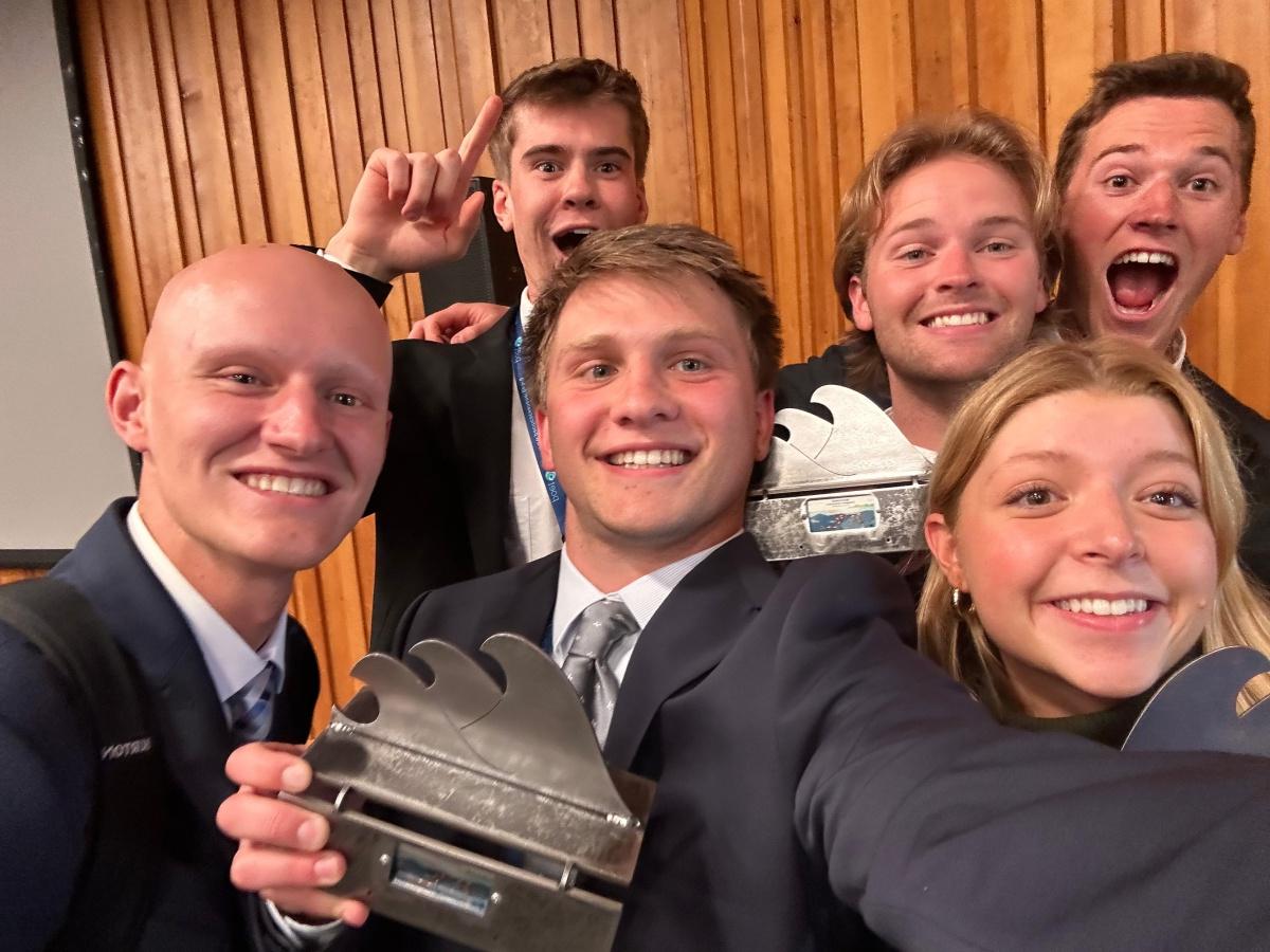 A selfie of six UNH students in suits holding wave-shaped trophies